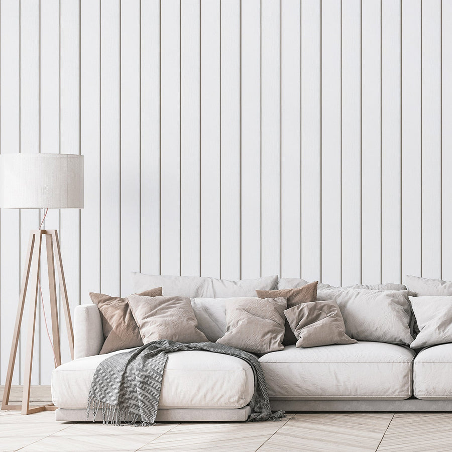 A living room with a floor lamp and a neutral coloured sofa featuring 'Airin' which is a white wallpaper featuring vertical wooden planks with realistic wood grain and subtle weathered texture in a rustic panelled design.