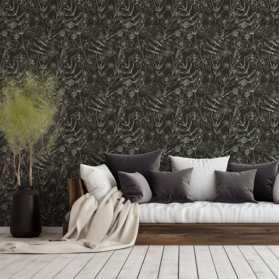 A living room with a leafy plant and low sofa scattered with pillows featuring 'Asa' which is a black and white botanical wallpaper featuring delicate outlined flowers, leaves and plant illustrations on a contrasting background in an intricate repeating pattern.