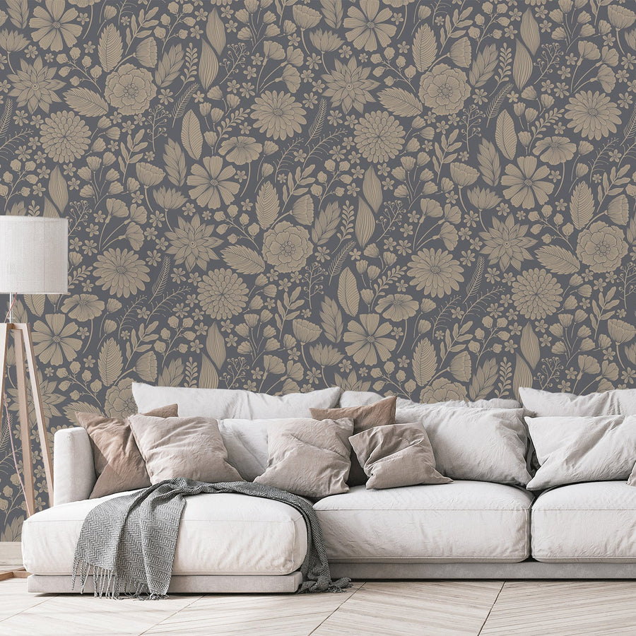 A living room scene with a beige sofa scattered with multiple cushions and a lamp showing 'Jessamine' which is a Blue botanical wallpaper featuring delicate floral blooms, leafy plants and fine botanical illustrations on a dark blue-grey background in a repeating pattern.