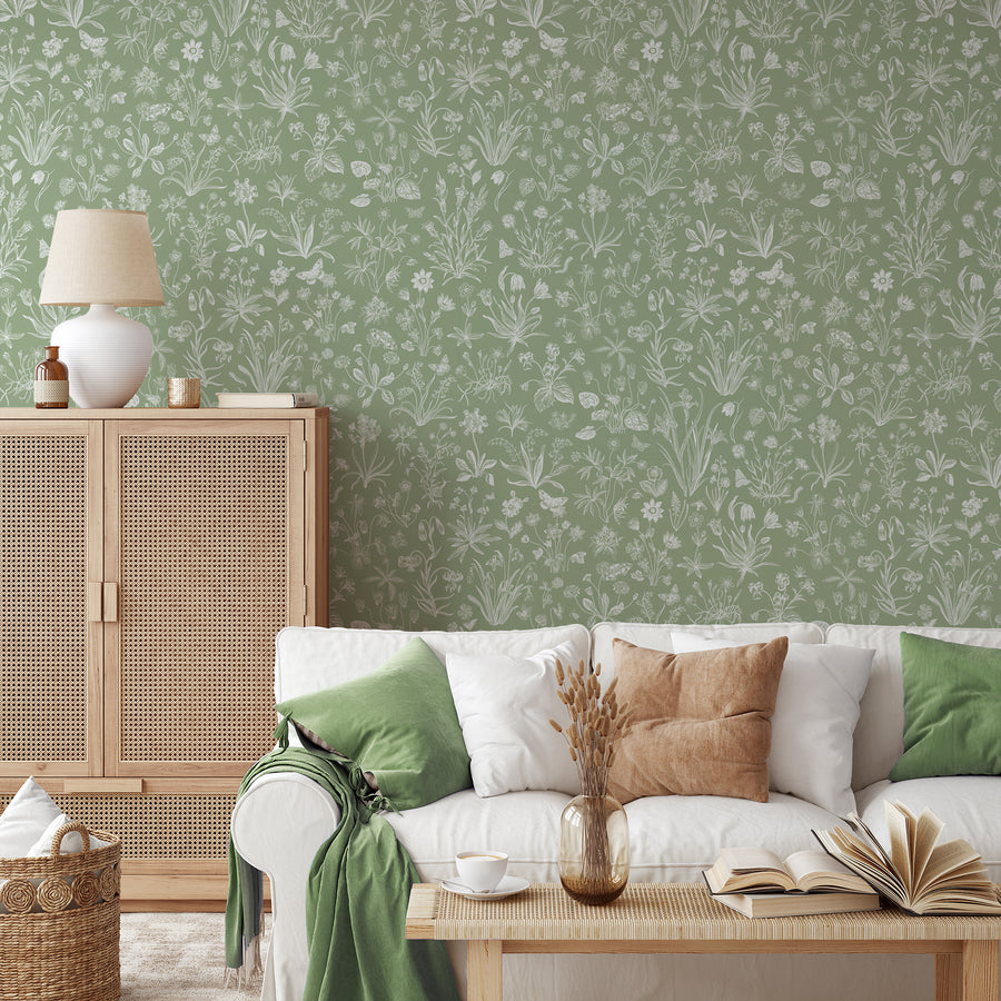 A soft, nature themed living room with rattan furniture featuring Peyton which is a green wallpaper with intricate white botanical flowers, foliage and butterflies arranged in a seamless repeating pattern.