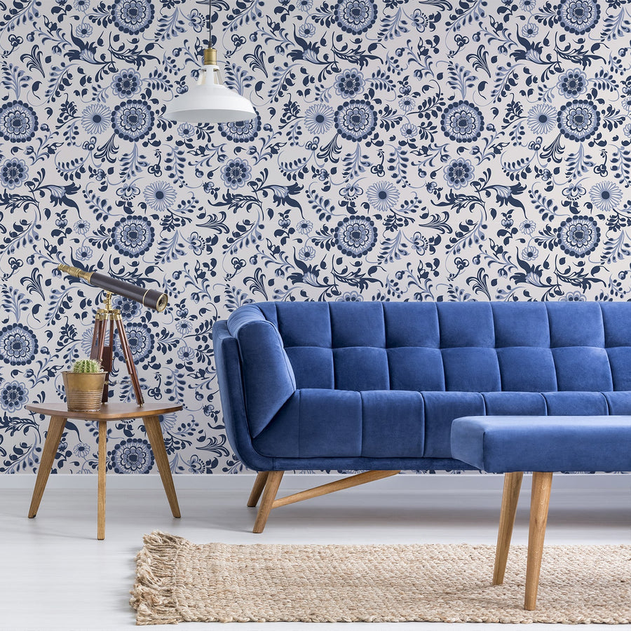 A living room with a velvet blue sofa, rattan rug and side table with a telescope and small catcus plant on top of it showing 'Bryony' which is a blue vintage wallpaper featuring navy floral blooms with leafy botanical details on a warm beige background in a classic repeating pattern.